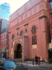 A five-story square building directly abuts a sidewalk. The facade is reddish brick, with two square windows on the second and three arched windows on the third floor. The main entrance juts forward from the facade, and is topped by an arch.