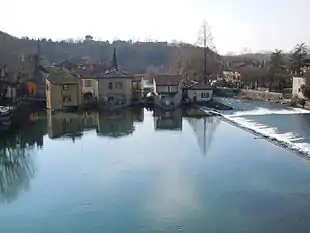 Photo of houses next to a river which flows past in the foreground and right. There is a high bluff on the left side.