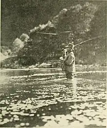 A man wearing waders, standing thigh deep in the river casting a flyrod