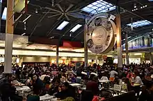 At bottom are numerous compactly-arranged tables filled with patrons eating. To the left is a large wooden support pillar, and to the right is a very large circular sign supported by a pillar at each edge; the centre of the sign contains the Vaughan Mills logo, which is surrounded on the periphery by six hand-drawn depictions of parts of the mall's interior, each separated by a small gold maple leaf. At top is a barrel-vaulted ceiling with one large skylight at its peak and two smaller skylights at its edge. Various fast food restaurants are seen in the background.