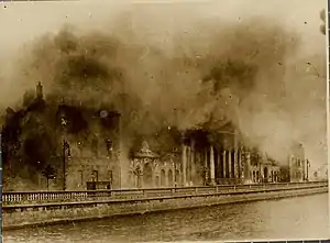 "Colourised photograph of an explosion above a number of buildings along a waterfront, with a thick pall of smoke obscuring the view of the tops of the buildings"