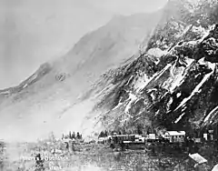 A small town shown at the base of a mountain. The mountain's face stands barren following a large rockslide and a light cloud of dust is visible in the air.