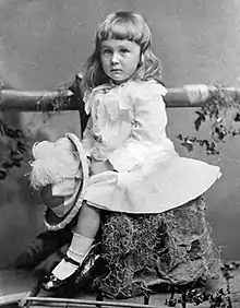 A young boy sits on a tree stump.  He holds a large feathered hat in his hand.