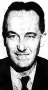 Black-and-white photographic portrait of a man with slicked-back hair, an angular smile (with possible tooth gap), faint mustache, and crooked nose. He wears a suit.