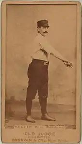 A baseball player is shown standing in his uniform, holding a baseball with his arm stretched out across his upper body.