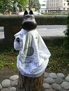 A dark metallic statue of Moomintroll wearing a white t-shirt with the Wikipedia logo and signatures in various languages.