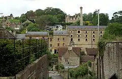 Yellow stone building with grey slate roof and grey chimney, surrounding by houses and trees. In the foreground is a path with a high stone wall and vegetation.