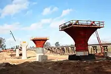 Several partially constructed vertical concrete supports rise from a dry riverbed on a sunny day