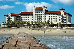 A stately white hotel building with a red-tile roof is seen from the end of a jetty extending from the beach.