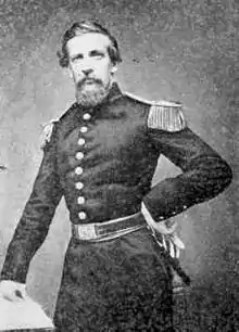 Black and white photo shows a bearded man standing with his left hand on his hip. He wears a dark military uniform with a single row of buttons and epaulettes.