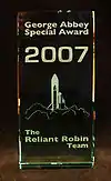 A 2007 George Abbey Award, stating in white text "George Abbey Special Award, 2007. The Reliant Robin Team." Depicted is a drawing of a rocket launching with a Reliant Robin attached to it.