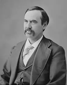 A man with dark hair and a mustache wearing a dark jacket and vest, light tie, and white shirt