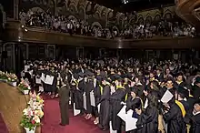 Young adults wearing ceremonial black robes and graduation caps stand at their seats in an ornate hall while onlookers in a three-sided balcony applaud above them.