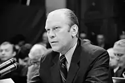 A man in a suit is seated at a table as he speaks into a bank of microphones. An audience is visible behind him.