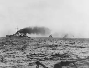 A line of large warships. Thick black smoke pours from their funnels as they steam through choppy seas.