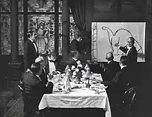 A black and white film still. A group of men sit around a dining table in the center. To the right, a man stands by and gestures at a large drawing of a dinosaur.