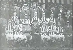 A football team comprising ten players in striped shirts and one in a shirt of a single colour pose for the camera. Five of the men are standing and the other six seated in front of them. Also posing with them are an elderly man in a bowler hat with a chain of office around his neck, and twelve men in business suits, some of whom are wearing hats.  A crowd of spectators is visible behind the group.