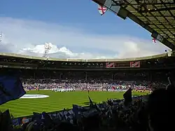 A stadium full of spectators. Those nearest the camera are waving blue and white flags.