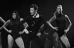 The grayscale picture of three people, two women and a man, who dance and put their hands on their hips. The man wears a black outfit, compound of a vest, a shirt and pants. The women wear similar leotards.