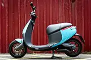 An electric motor scooter with a blue body, sitting on its kickstand in front of a red corrugated metal wall.