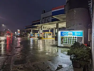 The Gogoro GoStation presents a bank of 20 or more green-handled batteries, brightly illuminated in an open shed on a rainy night next to a tiled petrol station in Taiwan.