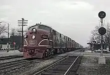 A red electric locomotive approaches the viewer from the left lane in a right-hand-drive context, opposite of the viewer. A telephone pole, stop sign, and railroad crossing can be seen in the distance.