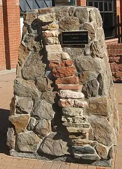 A stout pillar of motored irregular-shaped stone with insets of stacked more brick-shaped rock forming a column slanting to the right. A plaque on the pillar reads: "Grand Canyon Strata, Courtesy of Grand Canyon National Park".