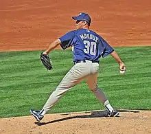 A man in a navy blue baseball jersey prepares to throw a baseball from a pitcher's mound with his right hand. His jersey reads "Maddux" in small tan print and "30" in larger tan print. He is wearing a navy blue baseball cap and gray baseball pants.