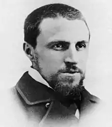 A black and white portrait photograph of a man with his hair cut very short and a goatee wearing a suit jacket with a high collar. He is looking ahead to his left, past the viewer