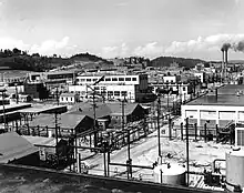 An array of industrial buildings with lots of power poles and wires, with a pair of smokestacks in the background