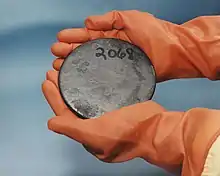 Two hands in brown gloves holding a blotched gray disk with a number 2068 hand-written on it