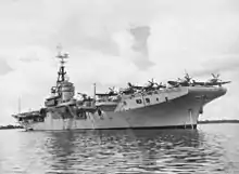 A aircraft carrier at anchor in still waters. Propeller aircraft are visible on her deck.