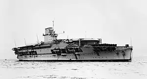 Right front view of a large anchored aircraft carrier with a flight deck that begins some ways back from the bow. Her island incorporates a funnel.