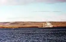 A warship at anchor. In the background is a flat, treeless landscape.