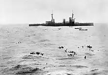 A large warship sits idly while several small boats pick up the numerous men swimming in the water.