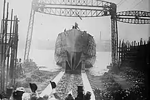 A black and white photograph showing a ship's hull with no superstructure sliding down a slipway into a waterway