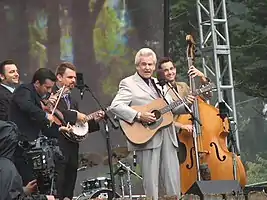 A man wearing a blue dress shirt, standing behind a microphone stand and holding a guitar backed by his band.