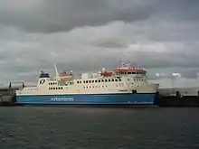 A large modern ferry with a blue hull and white topsides lies next to a harbour under grey skies.