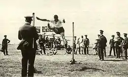 High jumper watched by a crowd of people, most wearing dark military uniforms with peaked caps