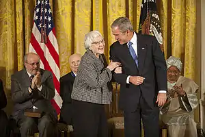 A color photograph of Harper Lee smiling and speaking to President George W. Bush while other seated Medal of Freedom recipients look on