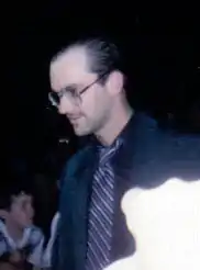 A man wearing a black suit jacket, a tie with narrow diagonal stripes, and eyeglasses. He has medium-length brown hair combed straight back and is slightly balding. A young boy can be seen in the background.