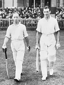 Two small men aged around 30 walk off the steps of a cricket ground pavilion onto the grass towards the centre of the playing area. Both wear light-coloured shirts, with rolled up sleeves, trousers and pads to protect their legs, all white. They wear batting gloves and hold a bat in readiness for play. The crowd sit behind a wooden fence; most are wearing suits and black cylindrical hats.