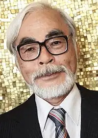 Close up photograph of Hayao Miyazaki, smiling and wearing a suit and tie in front of a gold-colored mosaic.