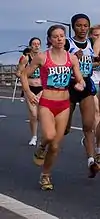 A woman in red runs alongside a group of athletes on a grey road.