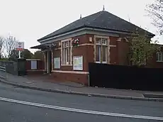 A small single-storey red brick building with cream stone window surrounds, mullions and transoms. An awning projects above the doorway and a slated pitched roof is topped with two finials.