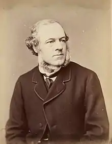 A seated man looks to the right of the viewer; he wears a suit and sports a neckbeard