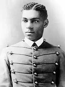 Cadet Henry O. Flipper in his West Point cadet uniform. It has three large round brass buttons left, middle and right showing five rows. The buttons are interconnected left to right and vice versa by decorative thread. He is wearing a starched white collar and no tie. He is a lighter colored African-American with plaited corn rows of neatly done hair. He is facing the camera and looking to the left of the viewer.