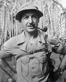 A standing man is smiling and staring off to his left. He has a smoke pipe in his mouth and is wearing a military uniform and hat.