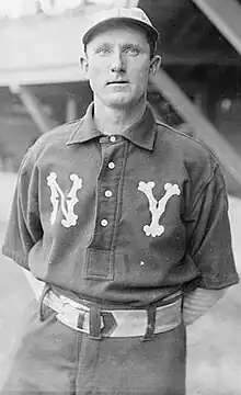 A baseball player is standing in his uniform, with his arm clasped behind his back.