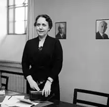 woman standing in an office with a window and photographs on the wall, behind a desk which has papers on it and her purse